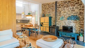 Uma sala de estar aconchegante com uma parede de pedra, lareira e televisão. Há uma área de jantar com mobiliário de madeira e uma cozinha ao fundo.