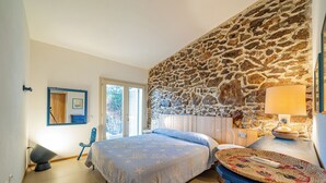 Quarto com parede de pedra, cama com colcha azul, mesa de cabeceira com abajur e vista para o exterior.