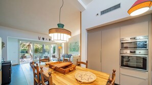 Cozinha moderna com duas fornos, área de jantar com mesa e cadeiras de madeira, e vista para o exterior através de grandes janelas.