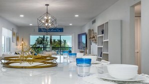 A modern living room with a central coffee table, a chandelier, and a view of palm trees through large windows.
