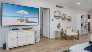 A living room with a large TV, a white cabinet with starfish designs, a round table with a decorative bowl, and a white armchair with a patterned pillow.