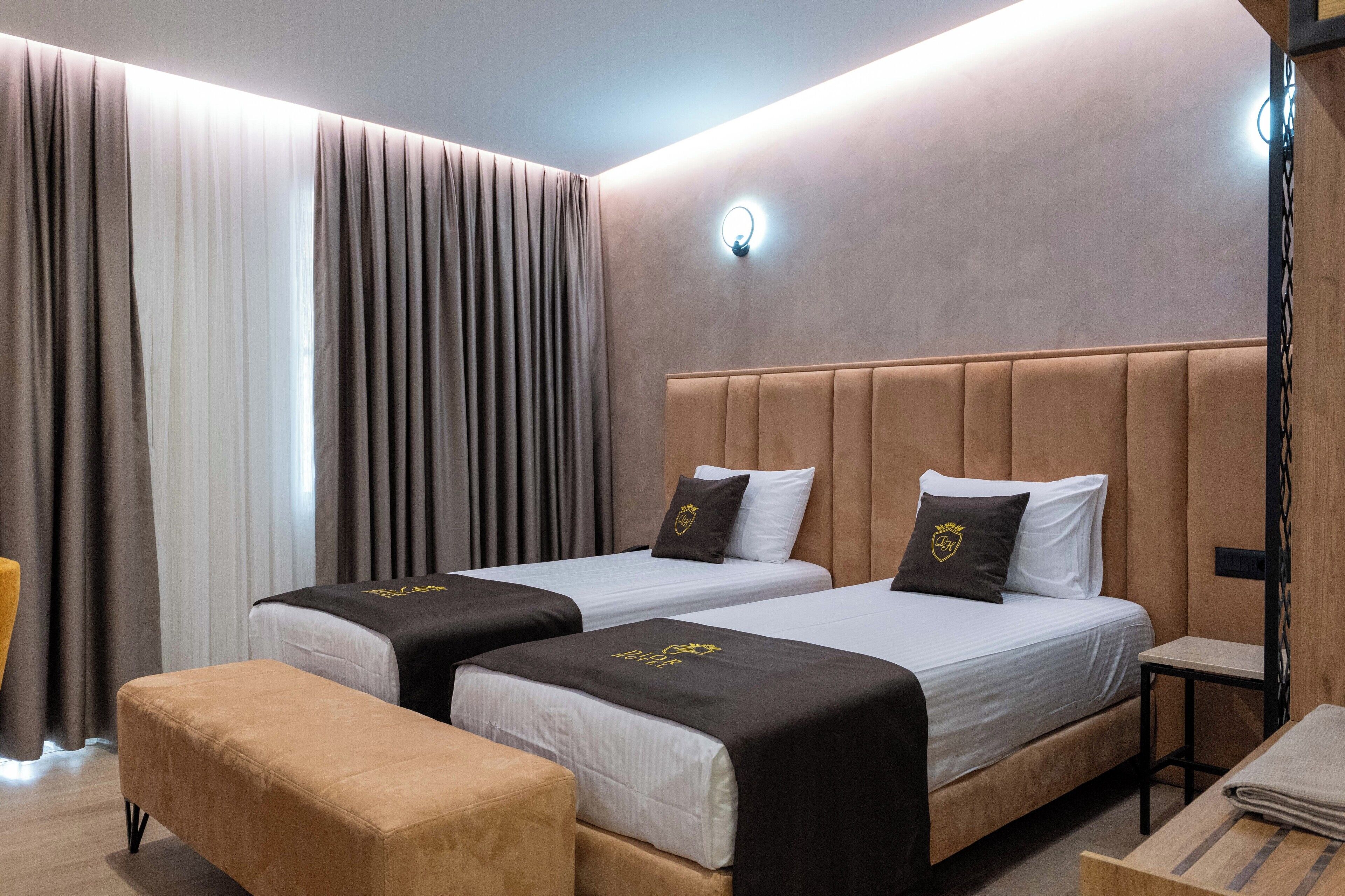 A hotel room with two beds, a bench, a small table, and a wall-mounted light.