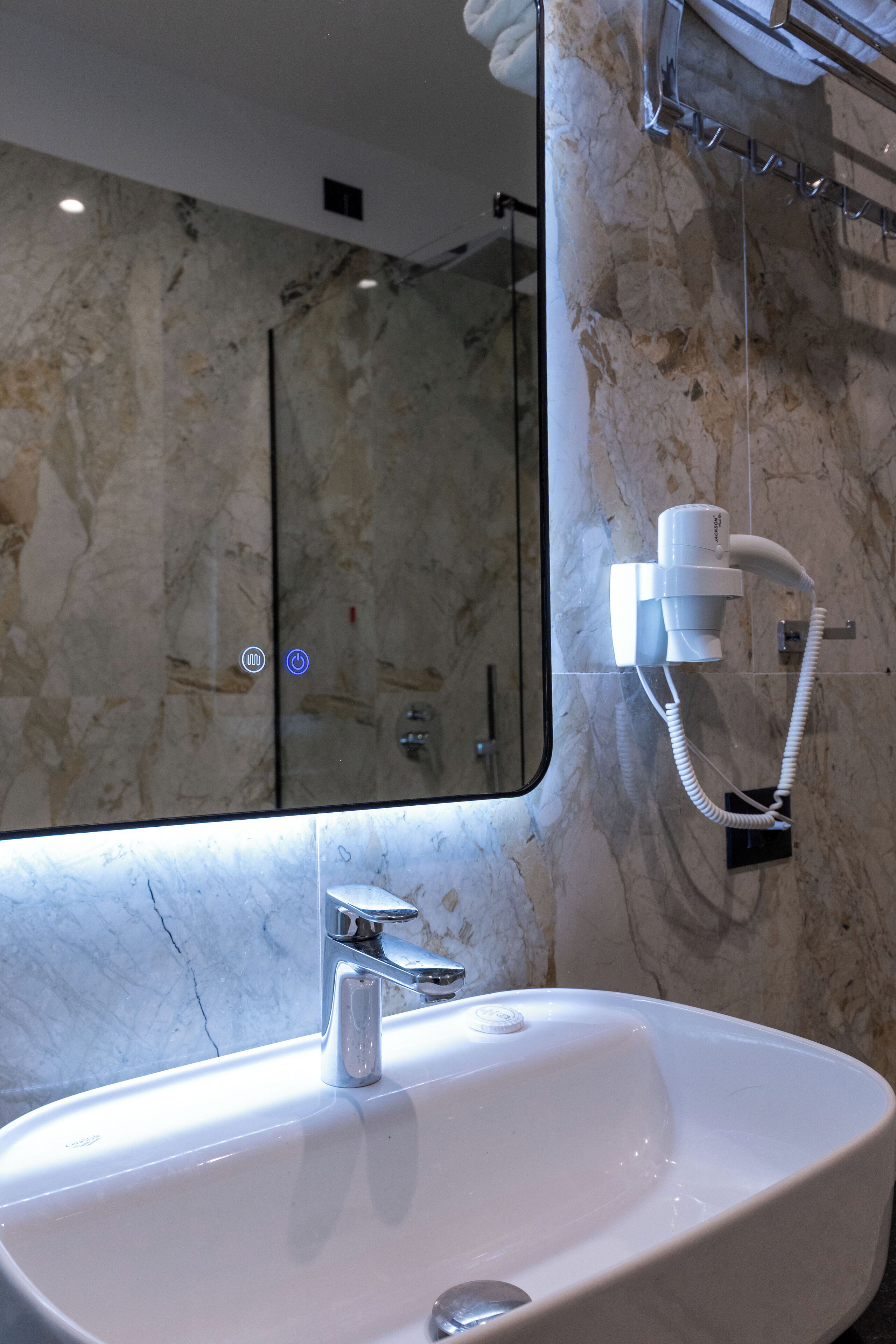 A bathroom with a marble wall, a large mirror, a white sink, and a hairdryer mounted on the wall.