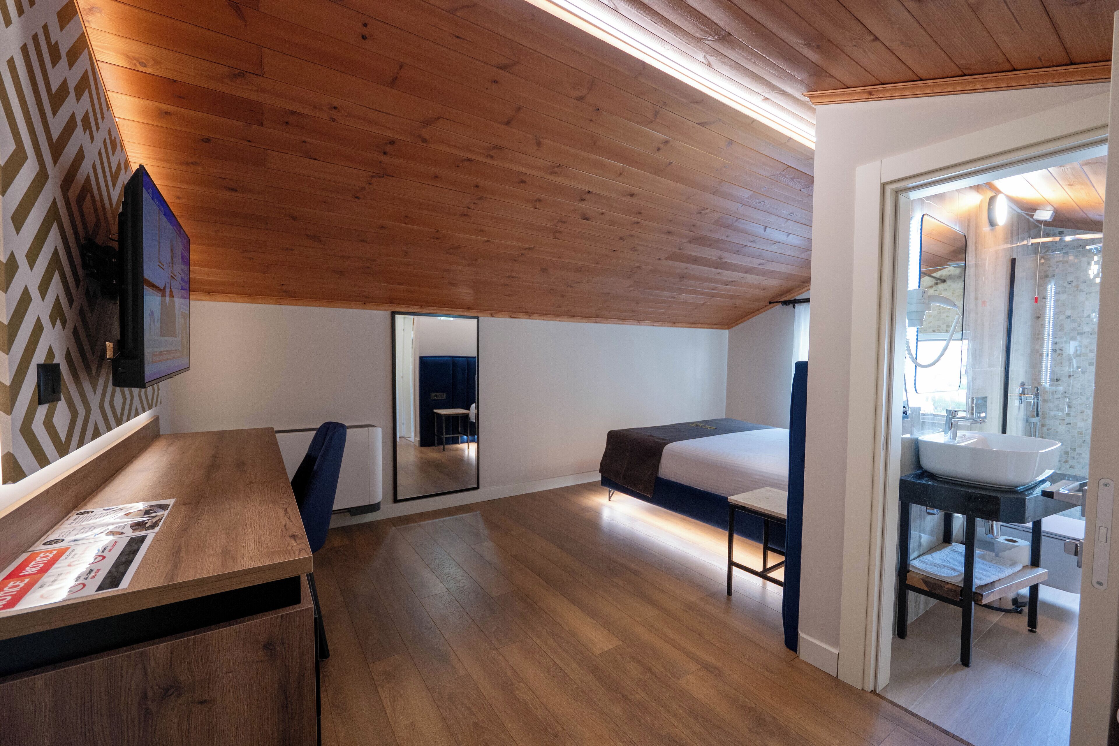 A modern bedroom with a wooden ceiling, a bed, a bedside table, a mirror, and a bathroom with a sink and mirror.
