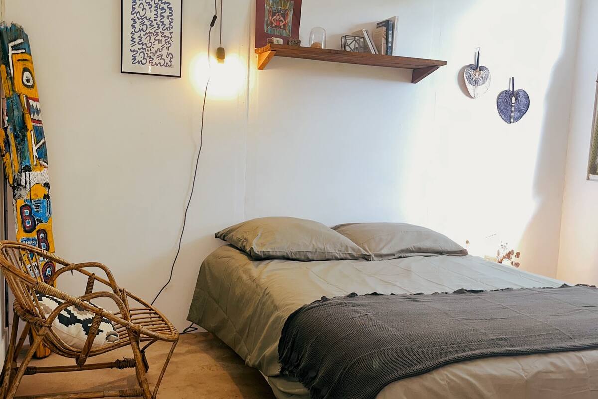 Un dormitorio con cama, una silla, un estante con libros y una lámpara en la pared.