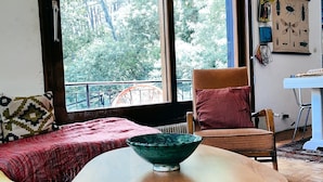 Een gezellige kamer met een groot raam, een houten tafel met een groene schaal, een rode bank en een rieten stoel.
