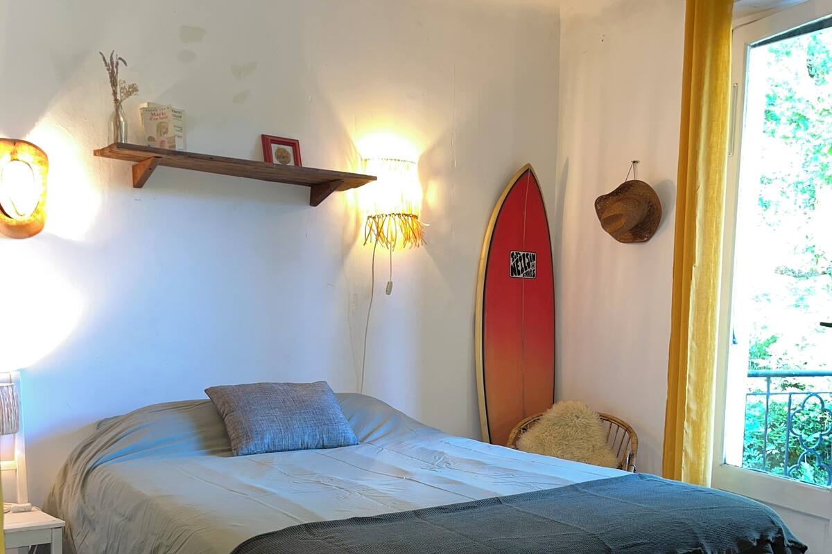 Un dormitorio con cama, una estantería con libros, una tabla de surf y una ventana con cortinas.