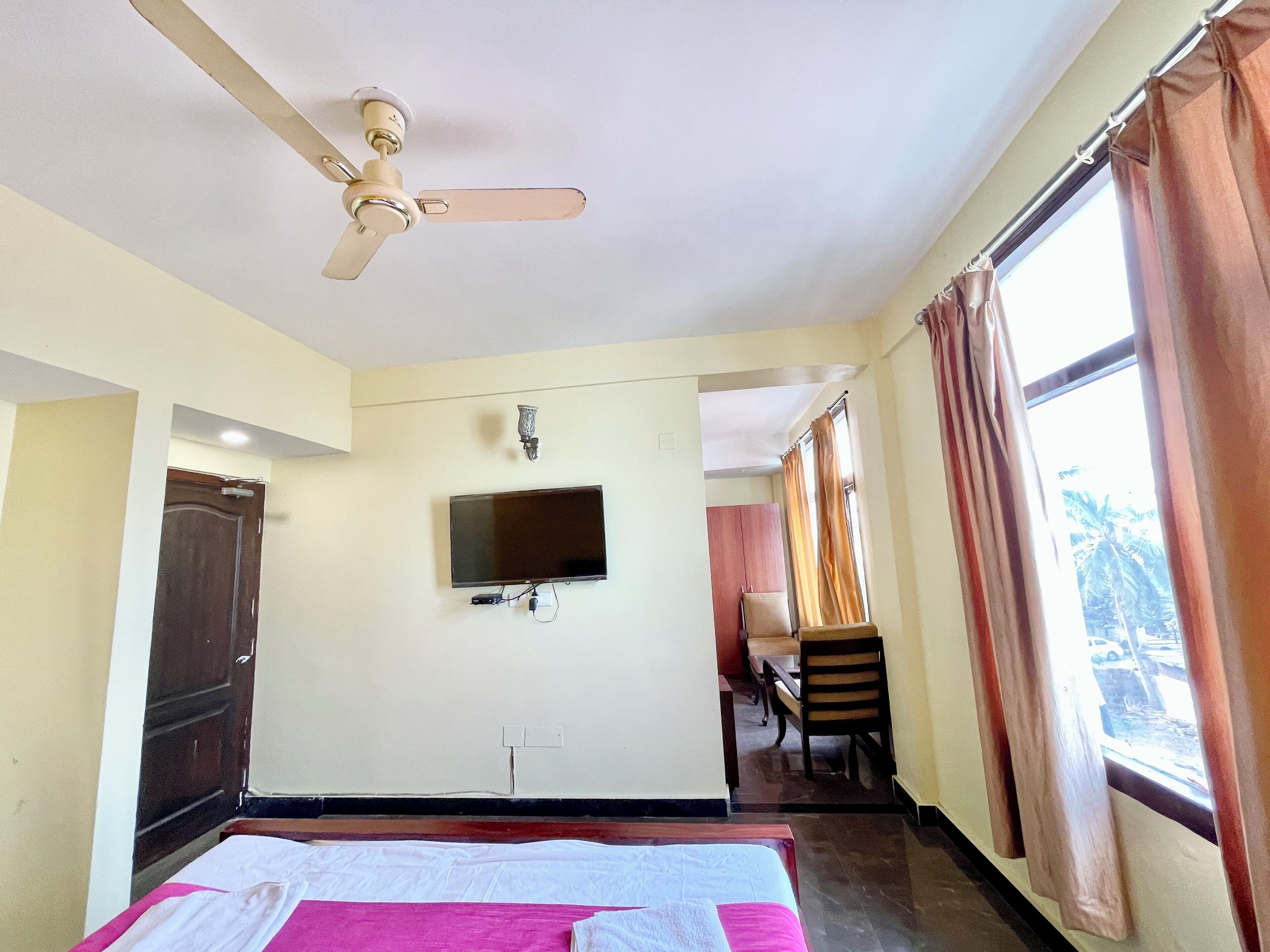 A hotel room with a bed, a ceiling fan, a flat-screen TV mounted on the wall, a window with curtains, and a door.