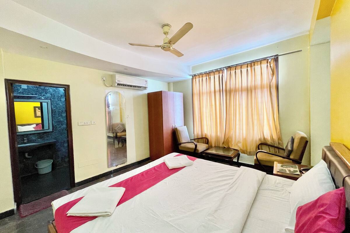 A hotel room with a bed, a desk, two chairs, a ceiling fan, an air conditioning unit, and a mirror.