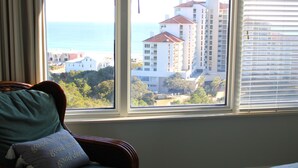 A room with a view of a coastal area, featuring a chair with a cushion and a window with blinds. - Summit at Topsail 1011 - 10th Floor (Miramar Beach)