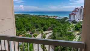 A view from a balcony overlooking a coastal area with dense greenery, a clear blue sky, and a distant view of a beach and buildings. - Summit at Topsail 1011 - 10th Floor (Miramar Beach)