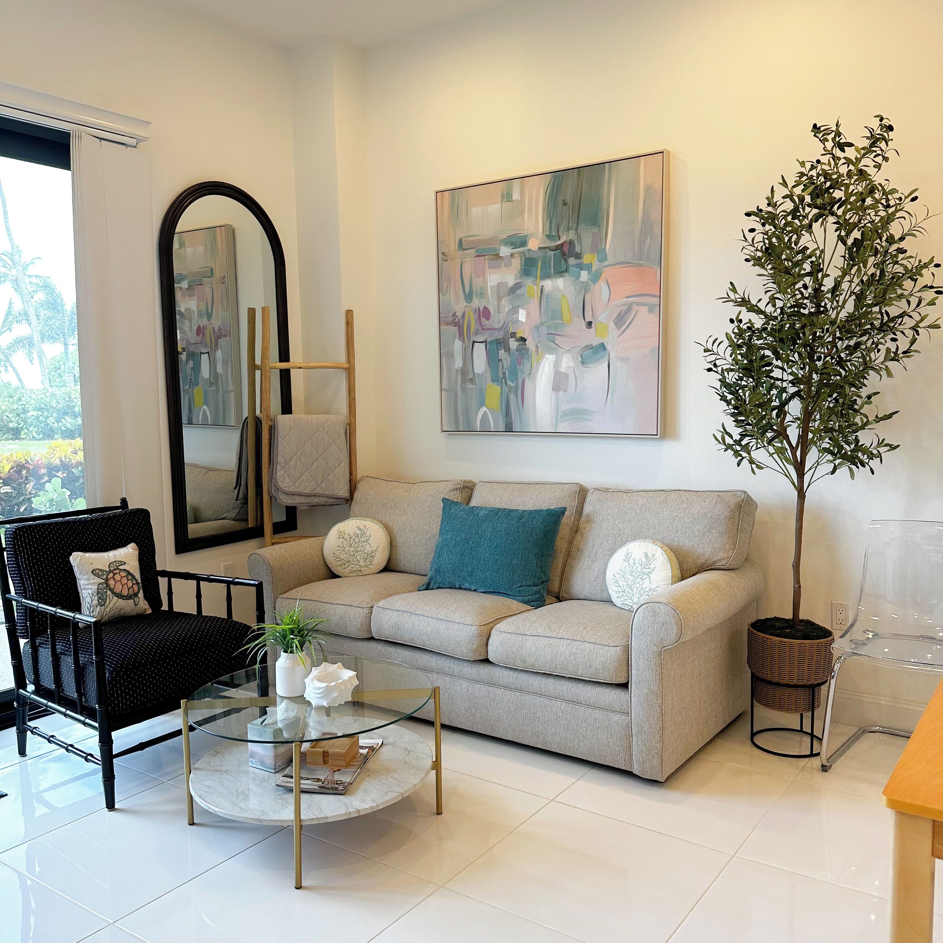 A modern living room with a beige sofa, a glass coffee table, a black armchair, a large abstract painting, and a tall potted plant.