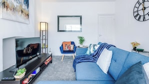 A modern living room with a flat-screen TV, a blue sofa, a small side table, a chair, and a large abstract painting on the wall. - Salford Oasis Retreat (Salford)