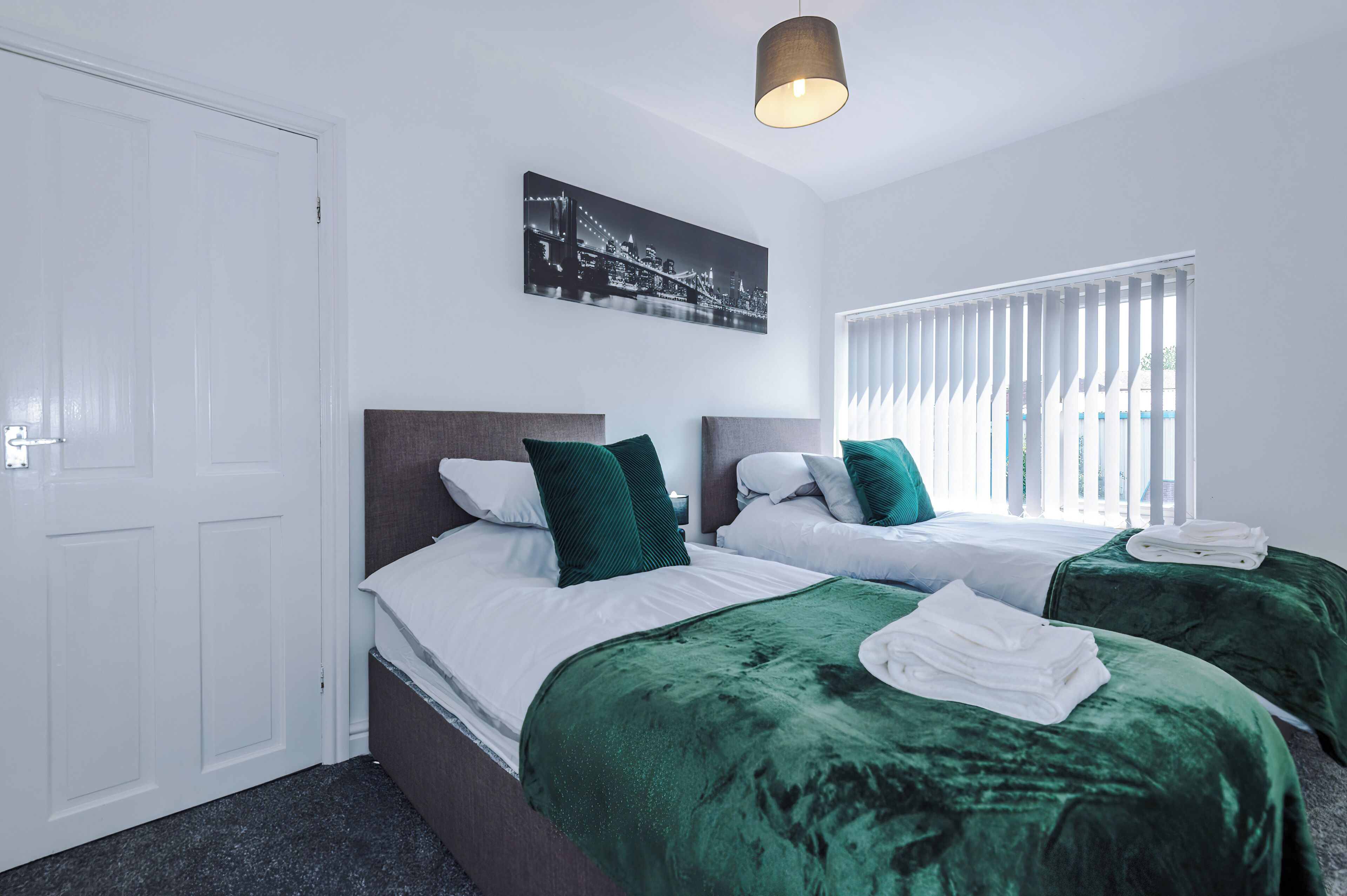 A hotel room with two beds, a cityscape artwork, a window with blinds, and a door.