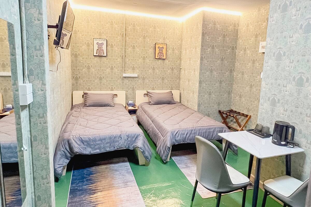 A small, compact room with two single beds, a small table with a chair, and a television mounted on the wall.
