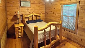 A wooden cabin bedroom with a bed, a nightstand, a lamp, a ceiling fan, and a window with blinds. - Cozy cabin on the water (Jasper)