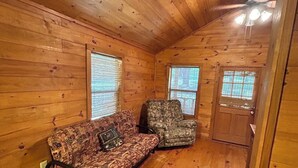 A cabin interior with wooden walls and floors, a patterned sofa, a camouflage chair, and a door with a window. - Cozy cabin on the water (Jasper)