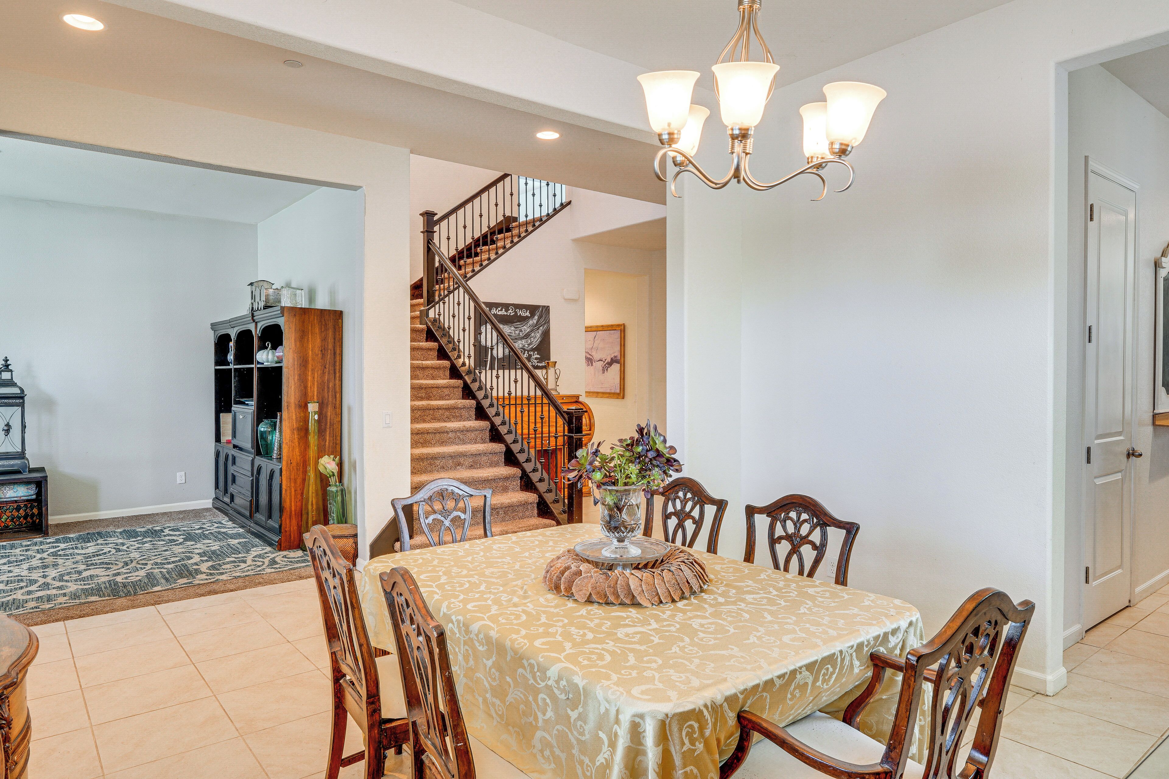 Un coin repas avec une table dressée pour quatre personnes, entourée de chaises en bois, d’un lustre et d’un escalier menant à un autre niveau.