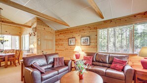 Un intérieur de chalet chaleureux avec deux canapés en cuir, une table à manger et une grande fenêtre équipée de stores.