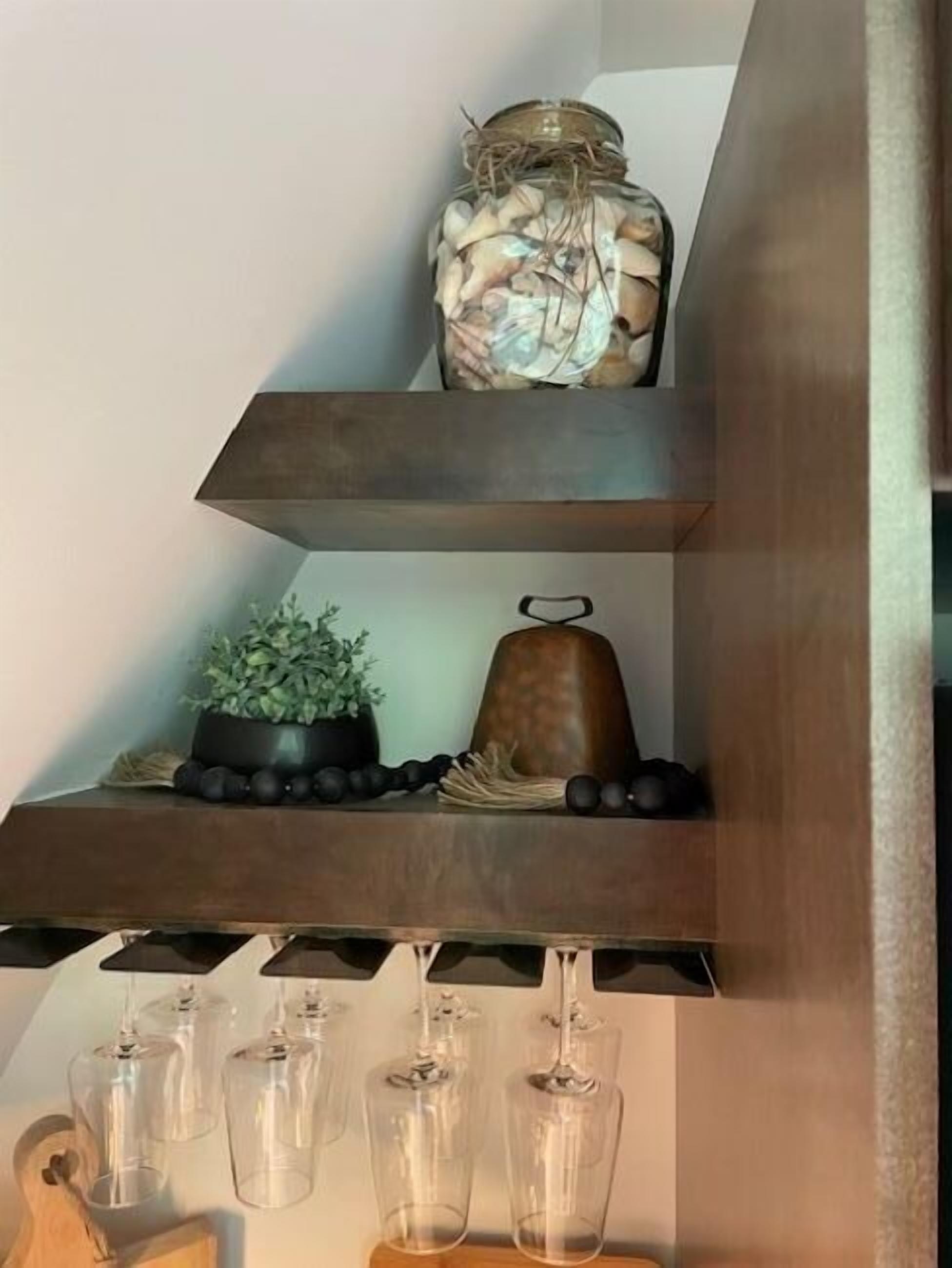 Una estantería de madera con un tarro de comida en conserva, una planta en maceta, una campana decorativa y copas de vino.