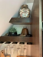 Una estantería de madera con un tarro de comida en conserva, una planta en maceta, una campana decorativa y copas de vino.