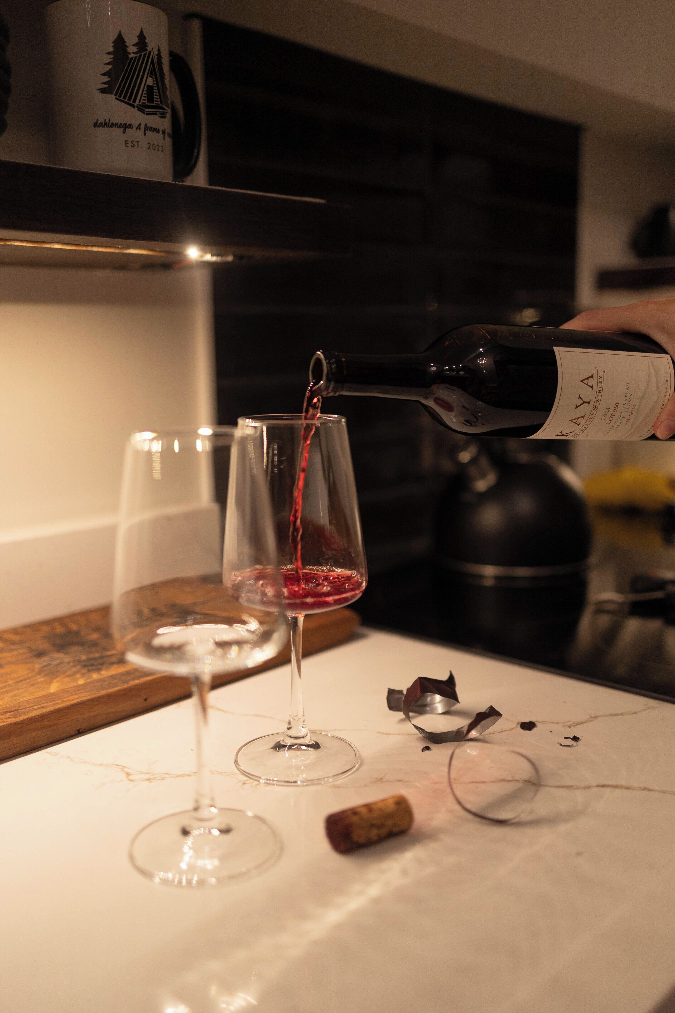 A wine bottle being poured into a glass, with another glass and a cork visible on a table.