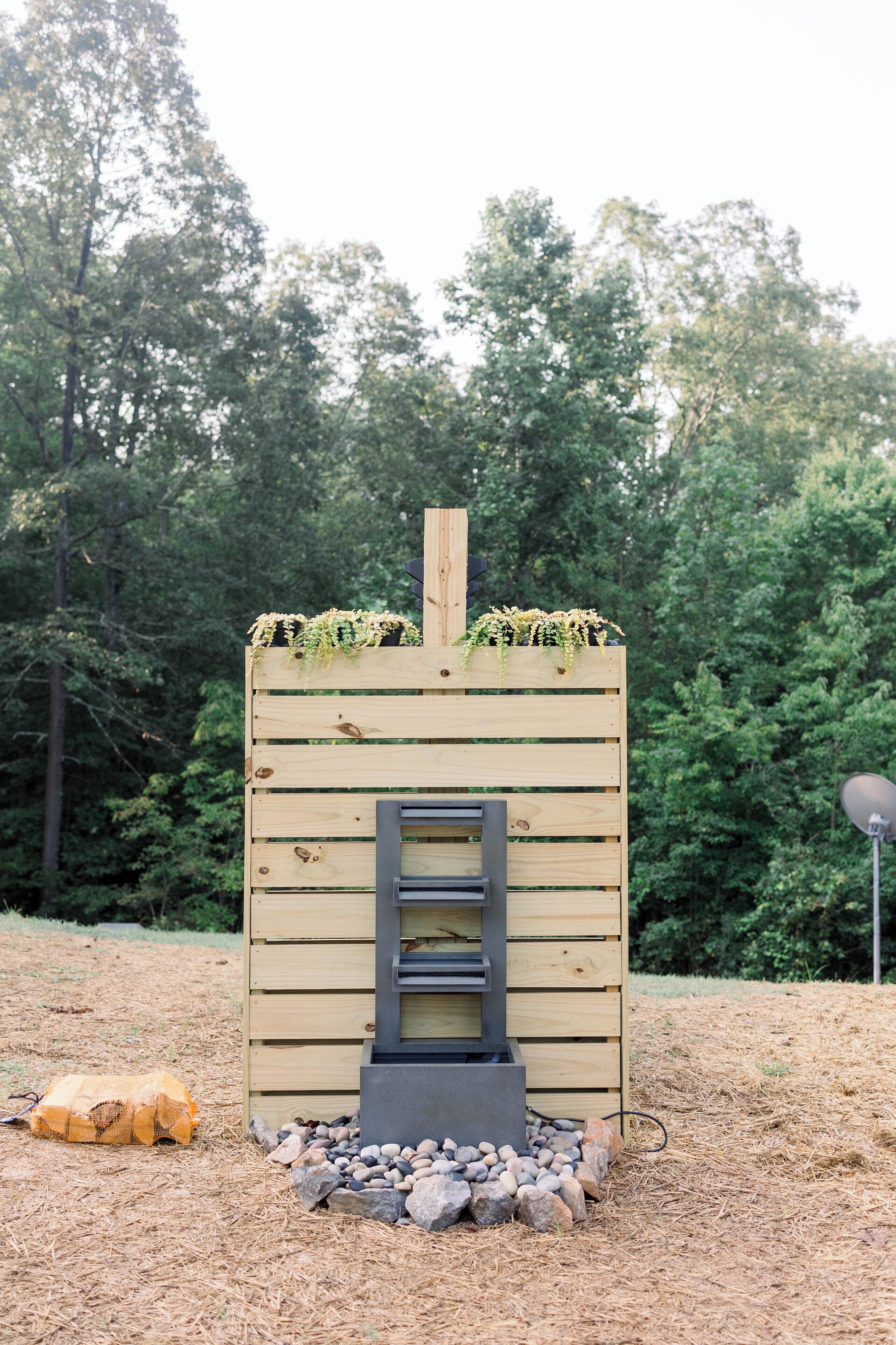 Una struttura in legno con una scala che conduce a una piattaforma, circondata da rocce e aghi di pino, con una vegetazione alberata sullo sfondo.