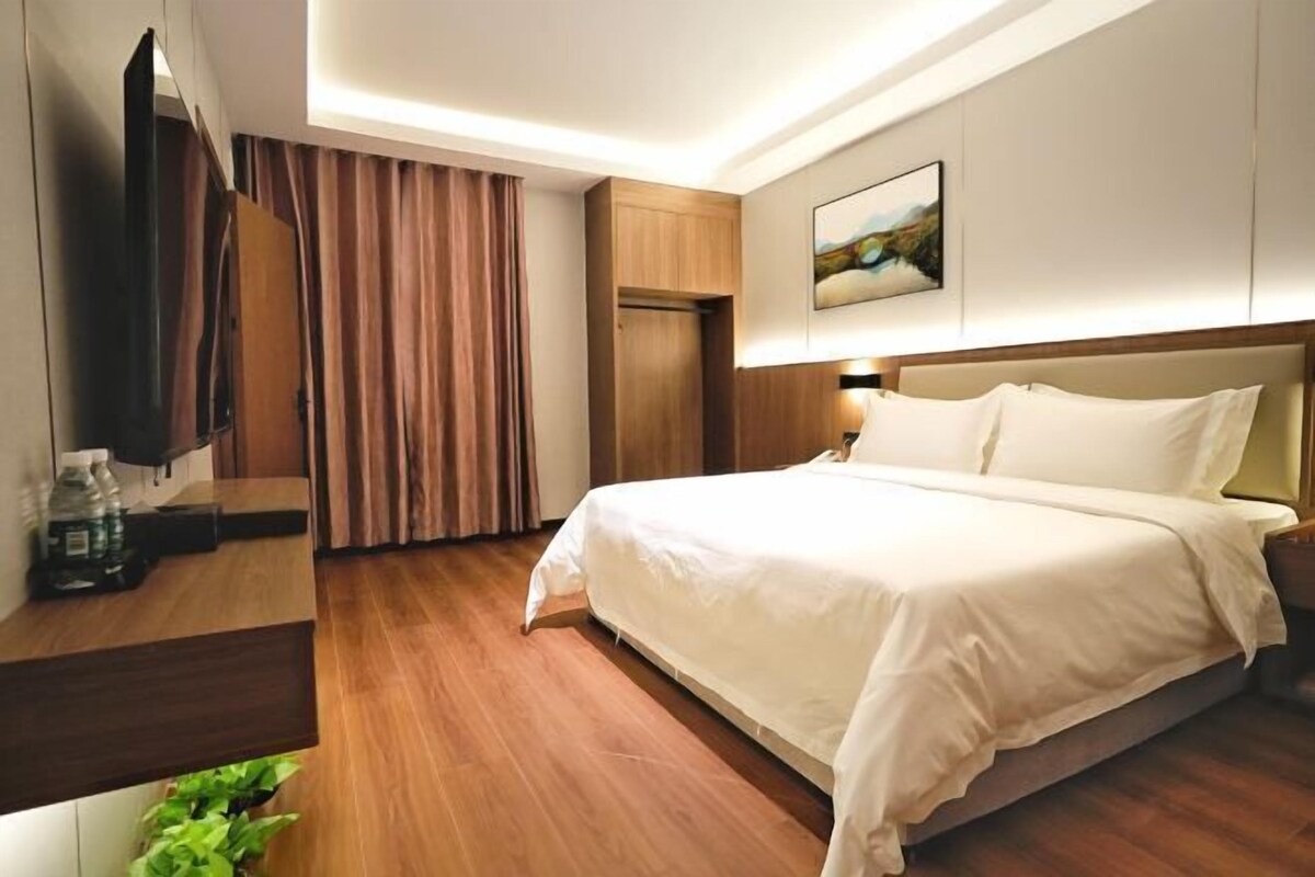 A hotel room with a bed, a TV, a wooden desk, a plant, and a painting on the wall.