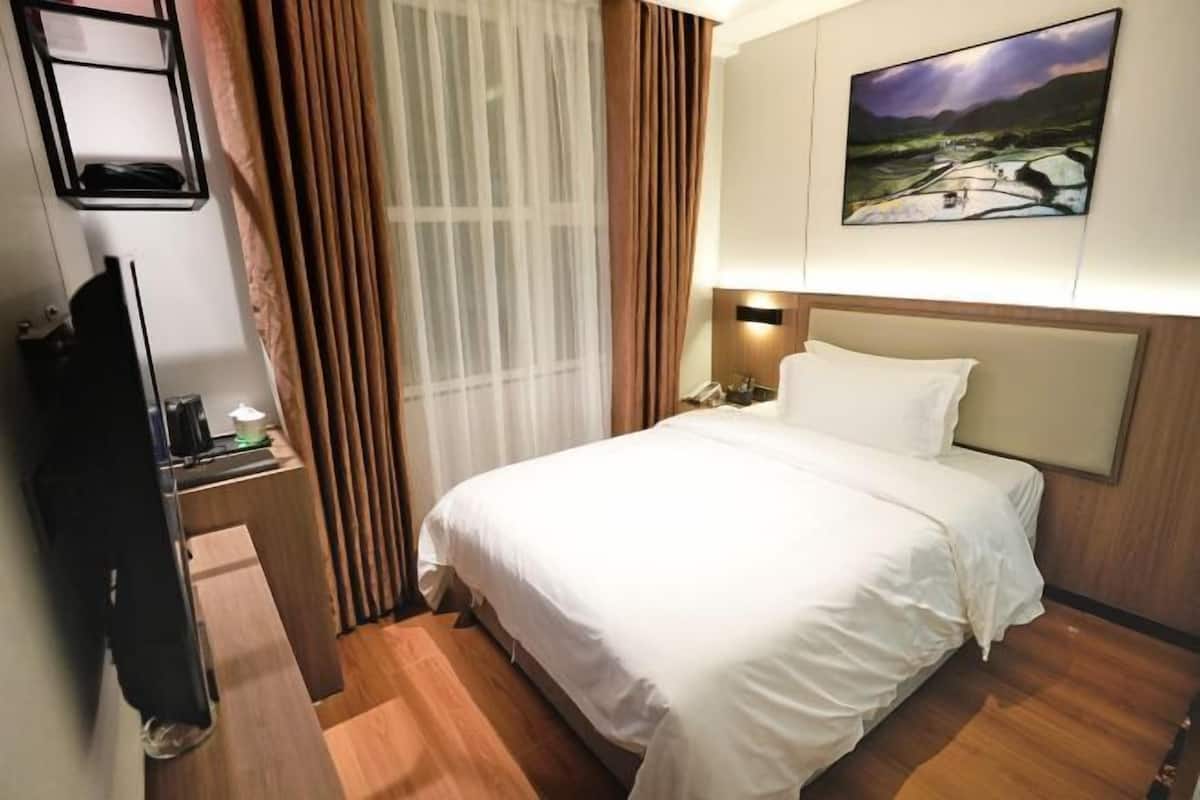 A hotel room with a bed, a nightstand, a lamp, a painting on the wall, and a window with curtains.