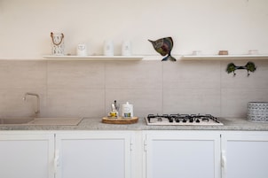 Una cocina con armarios blancos, encimera de granito, un lavabo, cocina a gas y diversos utensilios en los estantes.