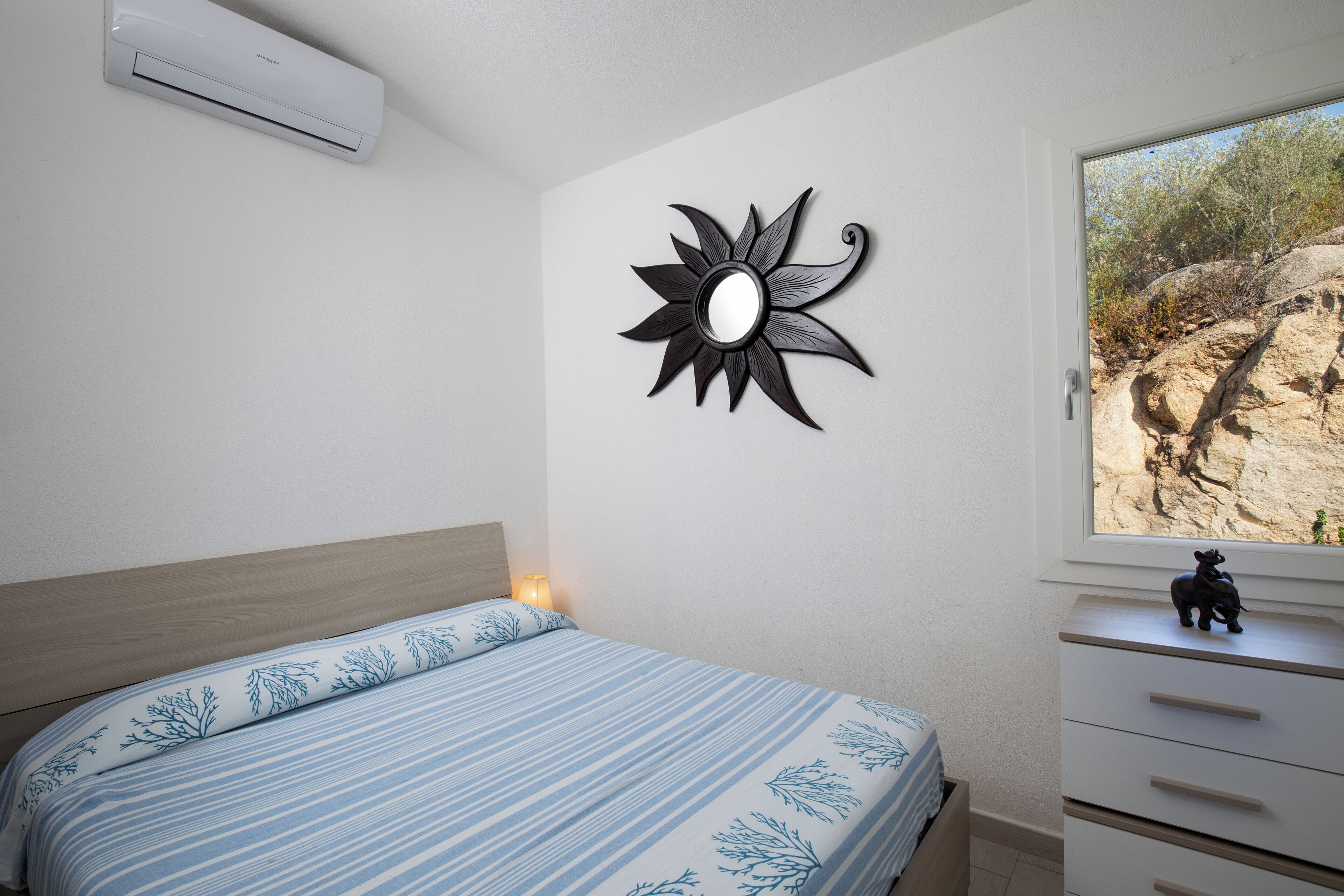 Un dormitorio con una cama, una mesita de noche, un espejo en la pared y una ventana con vistas a un paisaje rocoso.