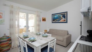Un'area pranzo con un tavolo bianco apparecchiato per quattro persone, un divano beige e una zona cucina con un piano cottura.