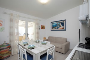 Un comedor con una mesa blanca dispuesta para cuatro personas, un sofá beige y una zona de cocina con estufa.
