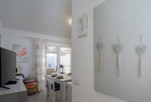 Uma sala de estar moderna com uma TV de tela plana, um espaço de jantar com uma mesa e cadeiras brancas, e uma janela grande com cortinas.