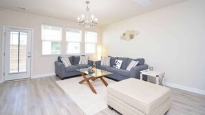 Un salon avec un ensemble de canapés gris, une table basse en verre, un pouf beige et un lustre.