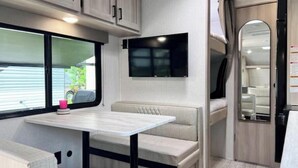 A compact RV interior with a dining area, a TV mounted on the wall, and a small seating area.