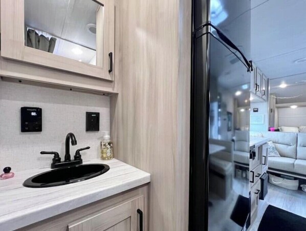 A modern kitchen with a black sink, white cabinets, and a glass door leading to a living area with a white sofa. - The Hoss Surf City, NC at Lanier's Campground (Hampstead)