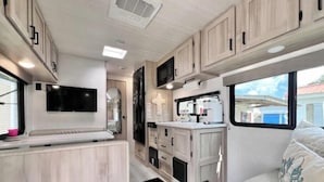 A compact RV interior with a kitchenette, a mounted TV, and a small seating area.
