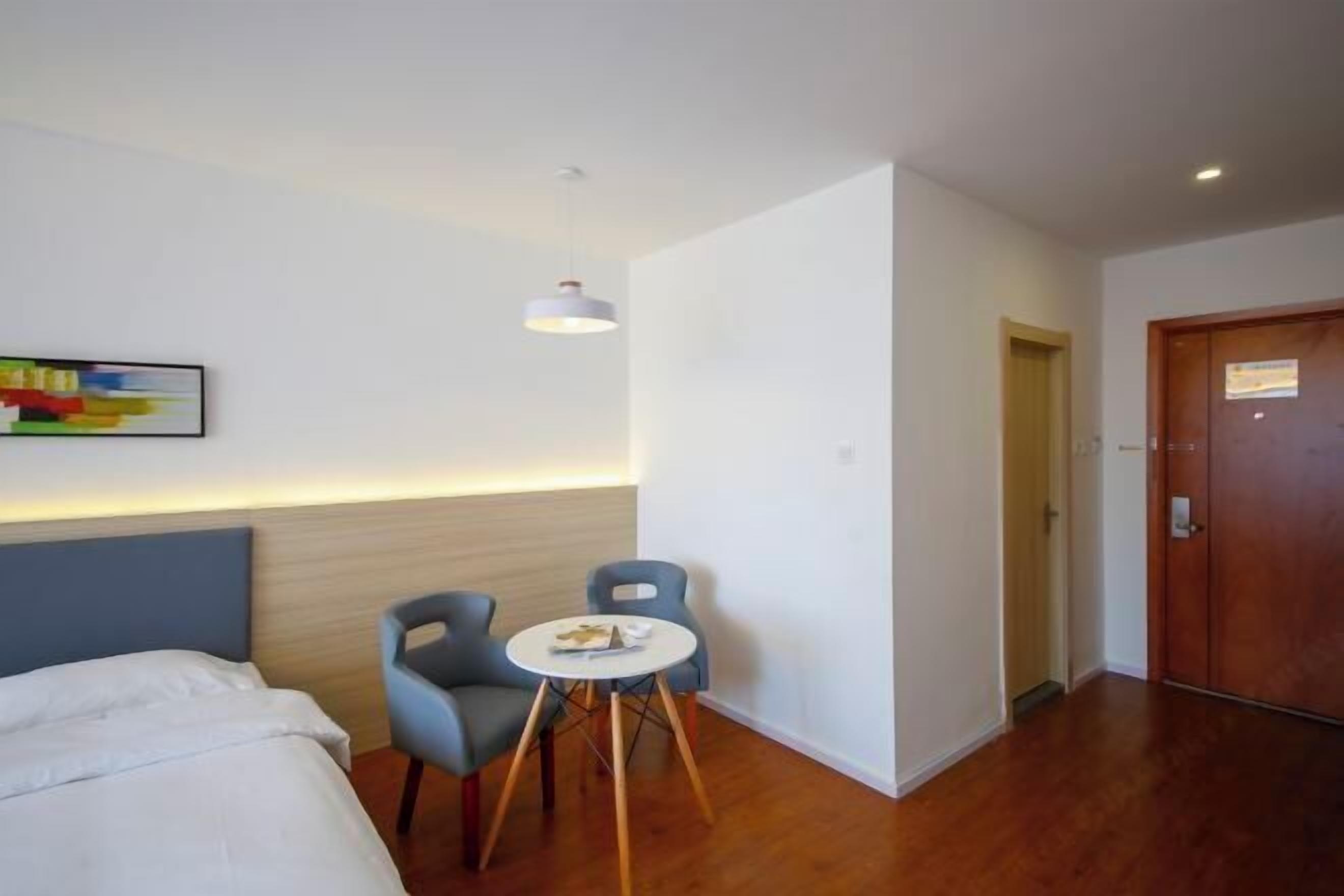 Quarto de hotel com cama, duas cadeiras, uma mesinha redonda, luminária pendente e porta de madeira.