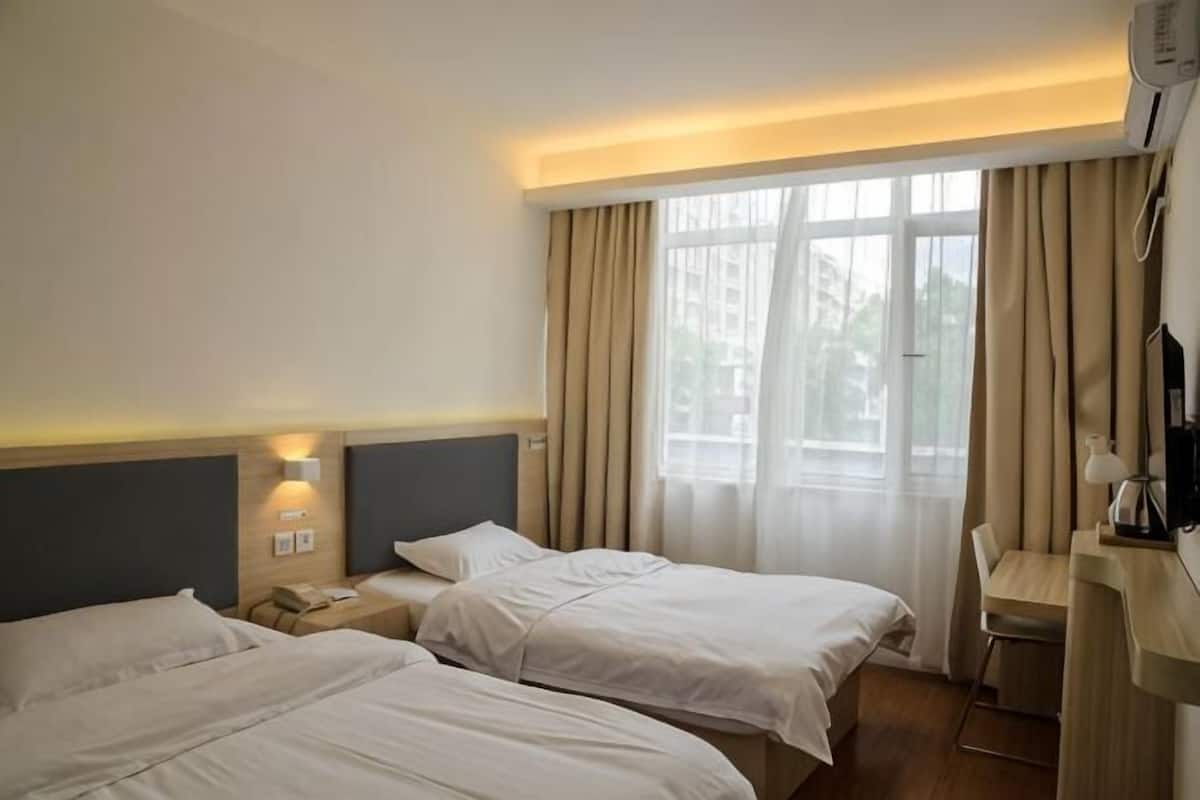 A hotel room with two beds, a desk, a chair, a window with curtains, and a wall-mounted phone.