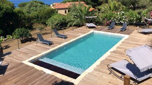 A rectangular pool with a wooden deck, surrounded by lounge chairs and greenery, overlooking a coastal area with buildings and trees. - 10 people sea view villa with pool in the south part of Corsica (Olmeto, Abbartelo)