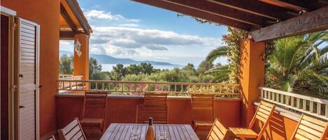 An outdoor patio with a wooden table set for two, a bottle of wine, and several glasses. The patio overlooks a lush landscape with trees and a distant mountain range.