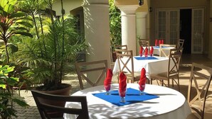 A dining area with round tables set for four, each with a white tablecloth and red napkins. There are potted plants and a columned structure in the background. - The Royal Hotel (San Fernando)