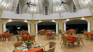 A spacious indoor dining area with multiple round tables covered in red tablecloths, surrounded by wicker chairs. - The Royal Hotel (San Fernando)
