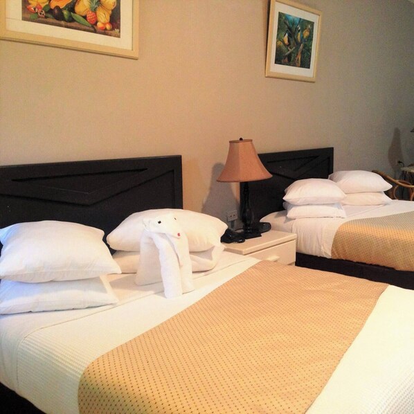 A hotel room with two beds, a nightstand with a lamp, and artwork on the walls. - The Royal Hotel (San Fernando)