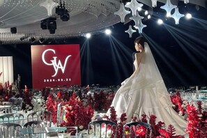 Una persona con un vestido blanco caminando sobre un escenario con un fondo rojo que muestra 'GW 2024.03.21'.