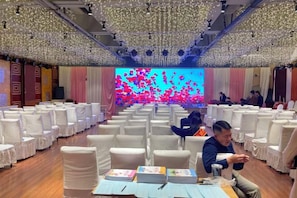 A banquet hall with rows of white chairs, a stage with a large screen, and a ceiling adorned with hanging lights.
