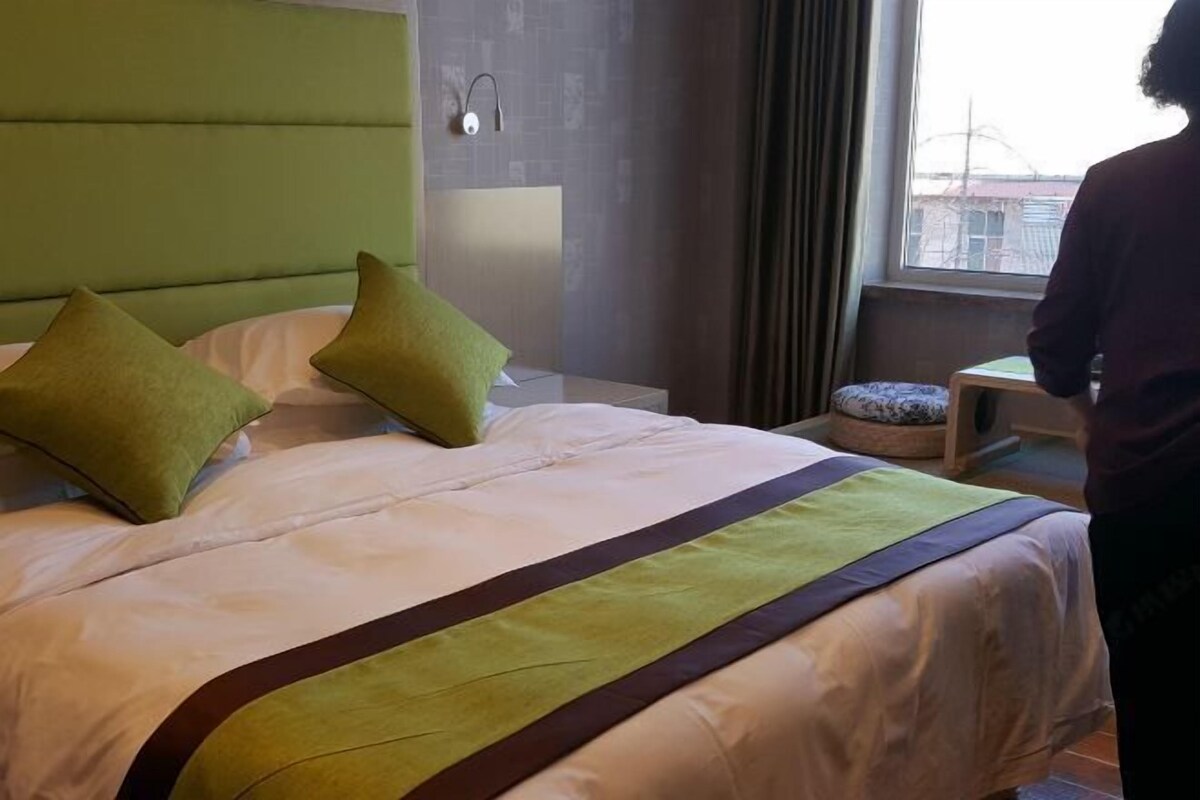 Quarto de hotel com uma cama, cabeceira verde, mesa de cabeceira e janela com cortinas.