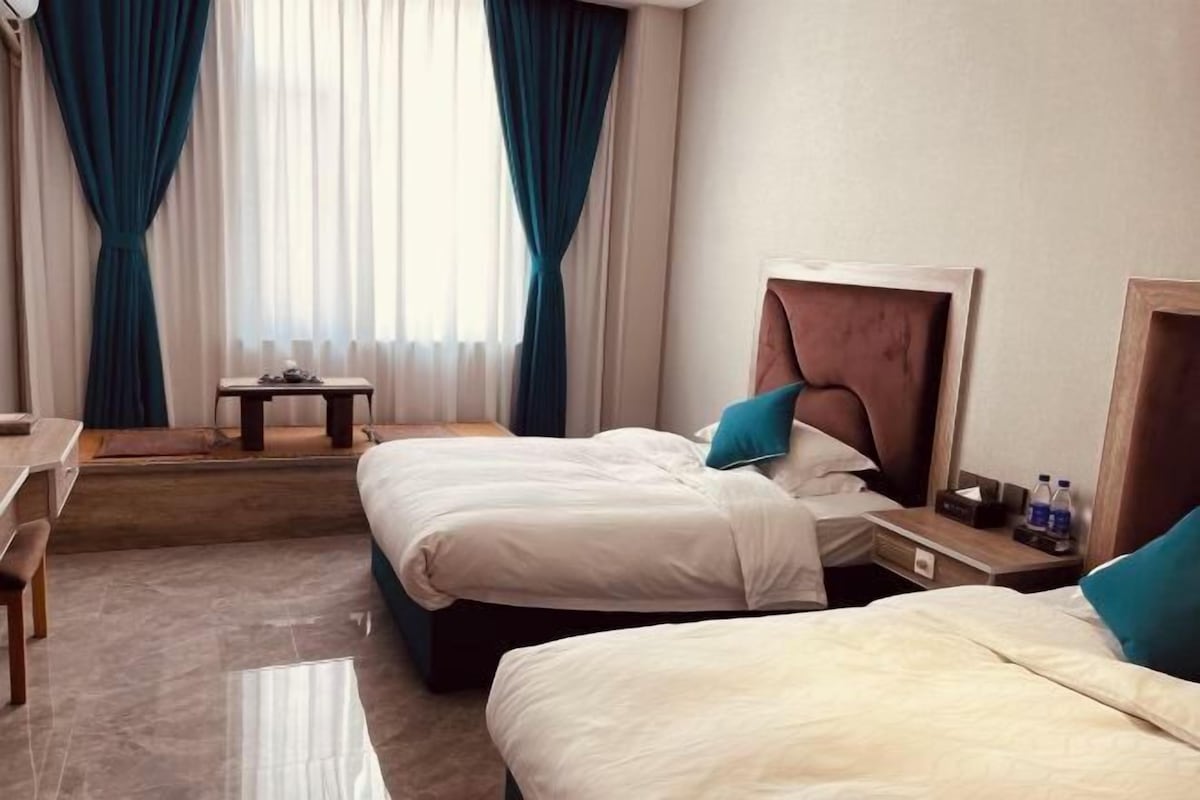 Quarto de hotel com duas camas, uma escrivaninha, uma cadeira, uma mesinha e uma janela com cortinas.