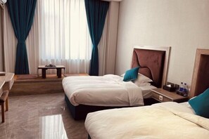 Quarto de hotel com duas camas, uma escrivaninha, uma cadeira, uma mesinha e uma janela com cortinas.